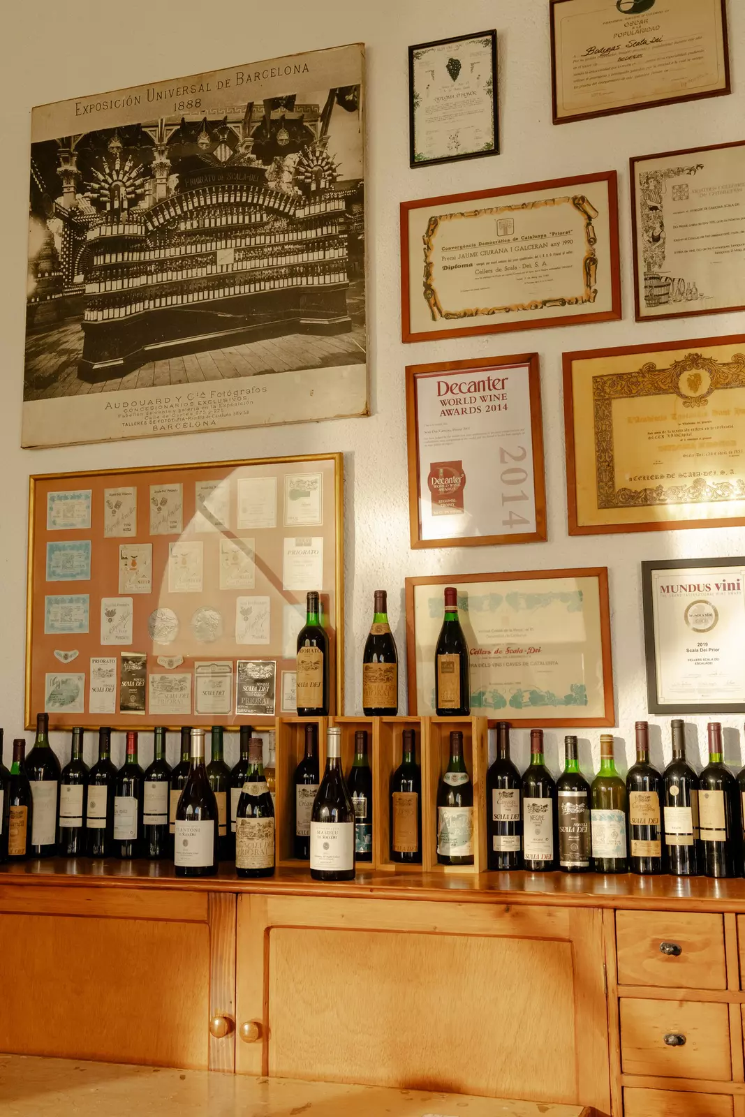 A wall with framed items and a shelf of bottles.