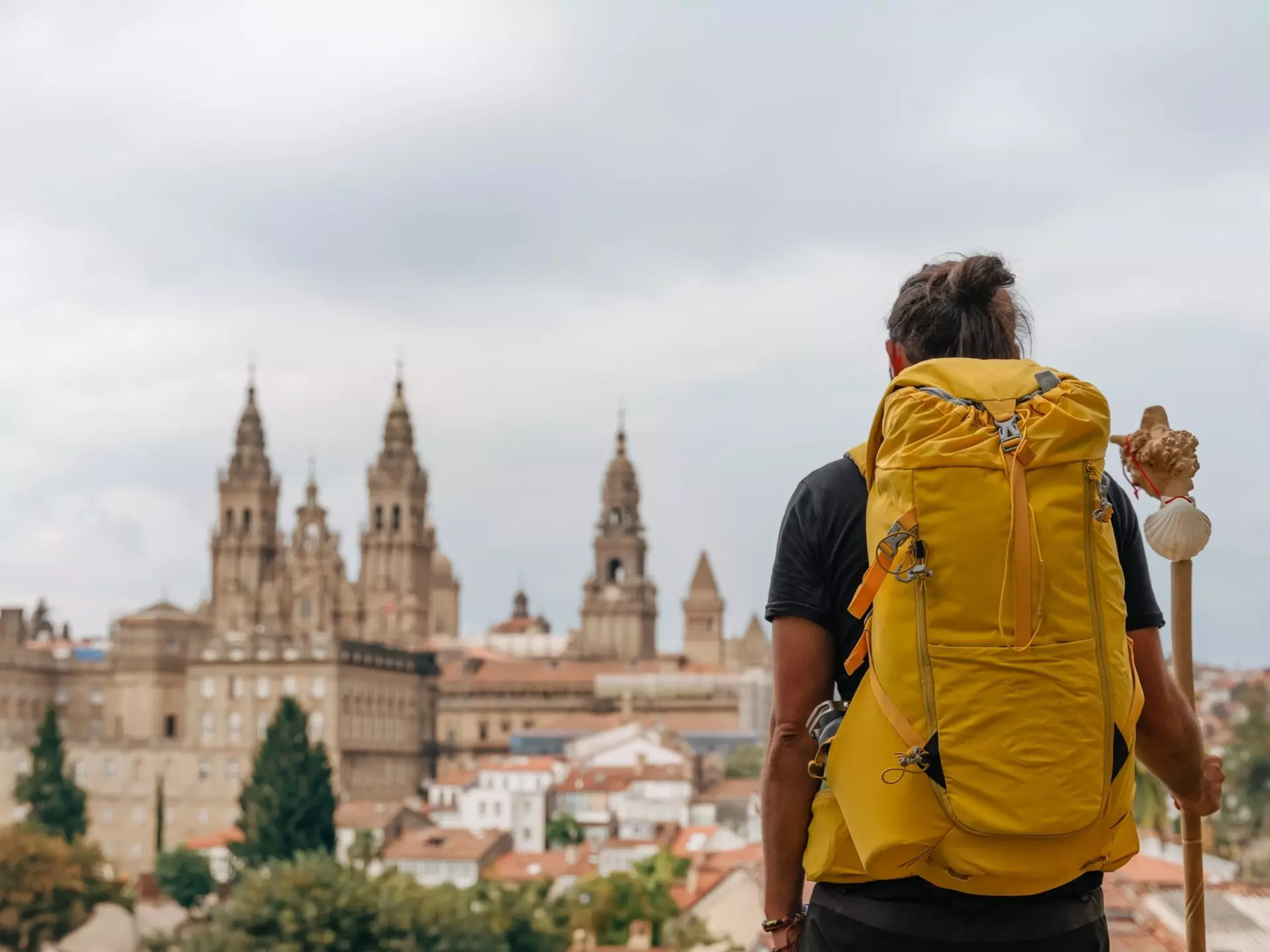 A hiker with a yellow backpack and a staff with a scallop shell looking at Santiago de Compostela in Spain.