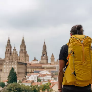 A hiker with a yellow backpack and a staff with a scallop shell looking at Santiago de Compostela in Spain.