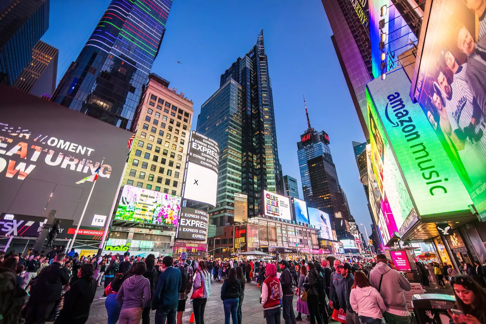 A crowd of people in Times Square as evening falls and neon signs light up the area.