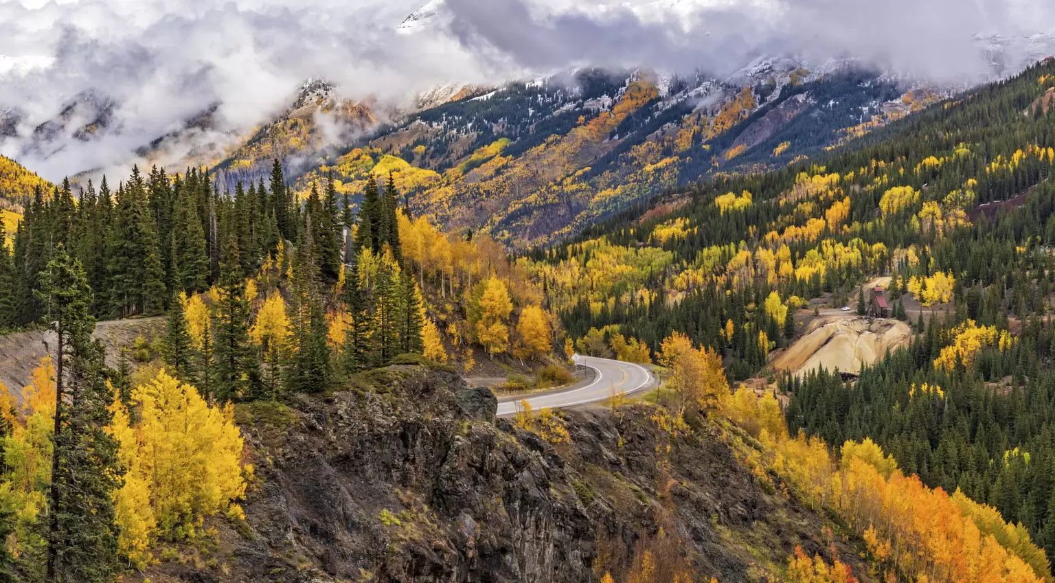 A road winds its way through mountains covered in trees colored yellow and green in autumn.