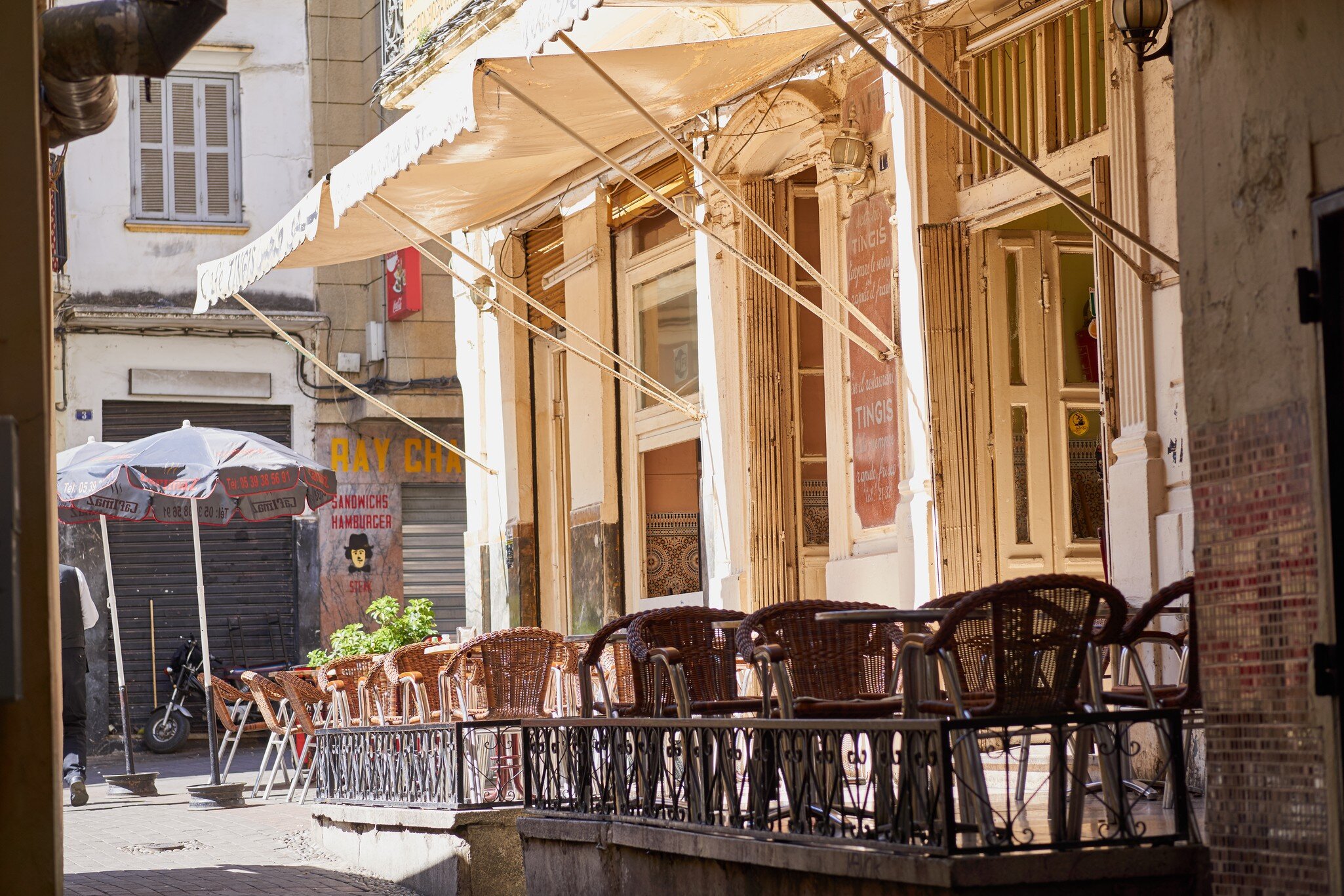 Empty wicker chairs sit behind an iron railing under a cream awning, with shuttered buildings and parasols along a lane