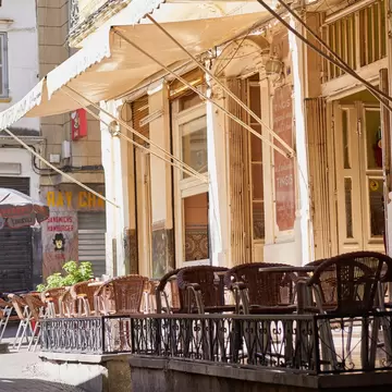 Empty wicker chairs sit behind an iron railing under a cream awning, with shuttered buildings and parasols along a lane