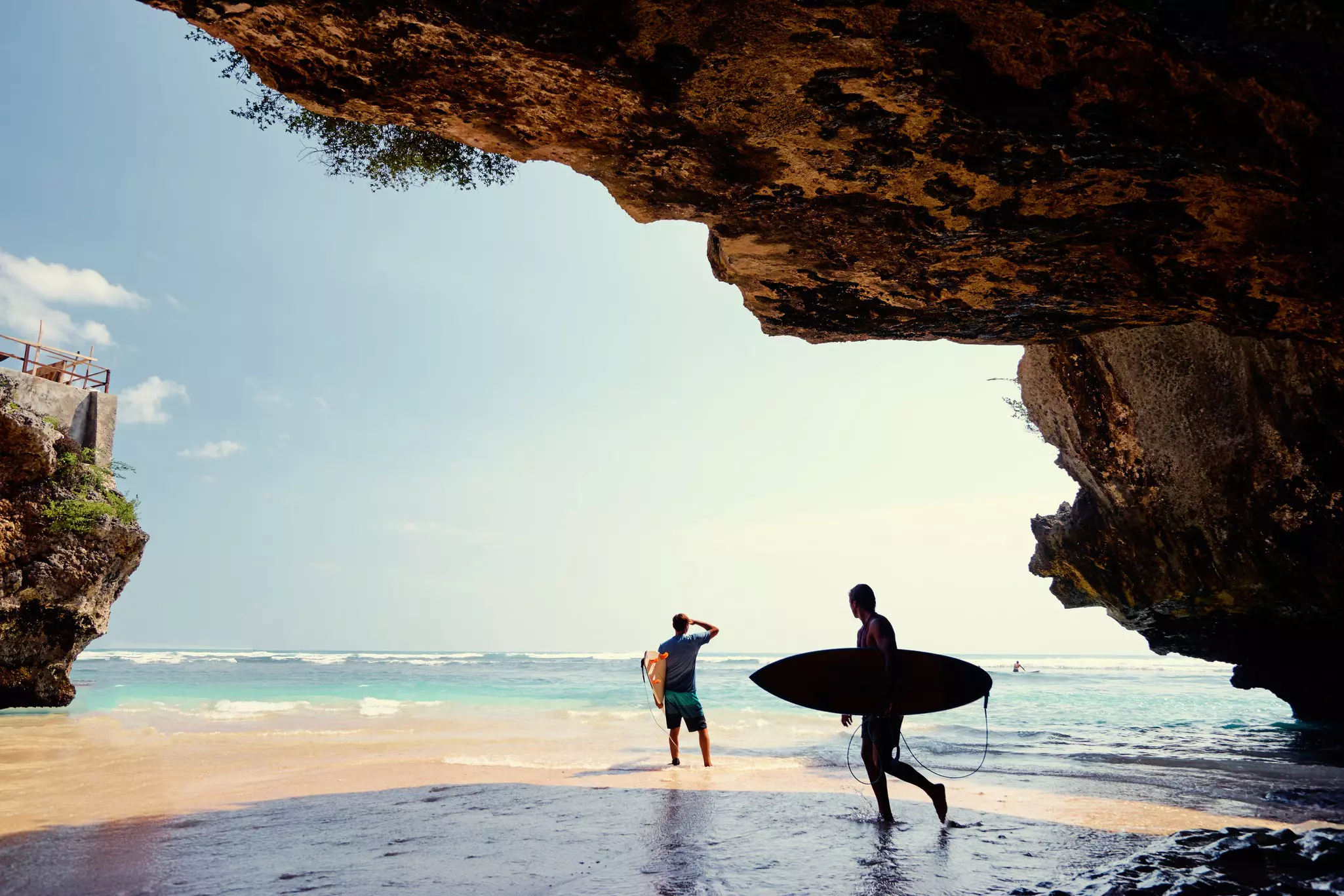 Surfers holding their boards are seen in silhouette as they stand on a beach in the shadow of a rock formation.