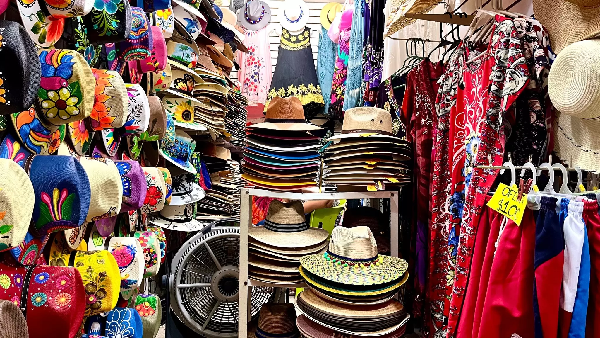 Straw hats, some brightly colored, and colorful clothing for sale in a crowded space.