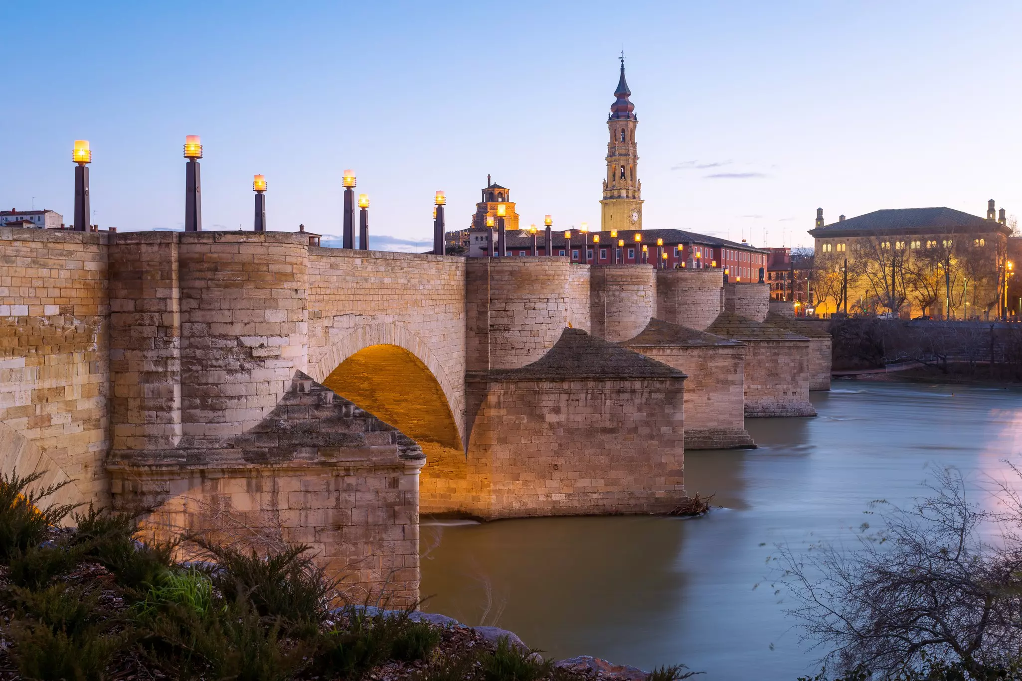 A view of the Puente de Piedra in Zaragoza, Aragon, Spain in the dusk light with the River Ebro flowing under it. Lights are on along the bridge.