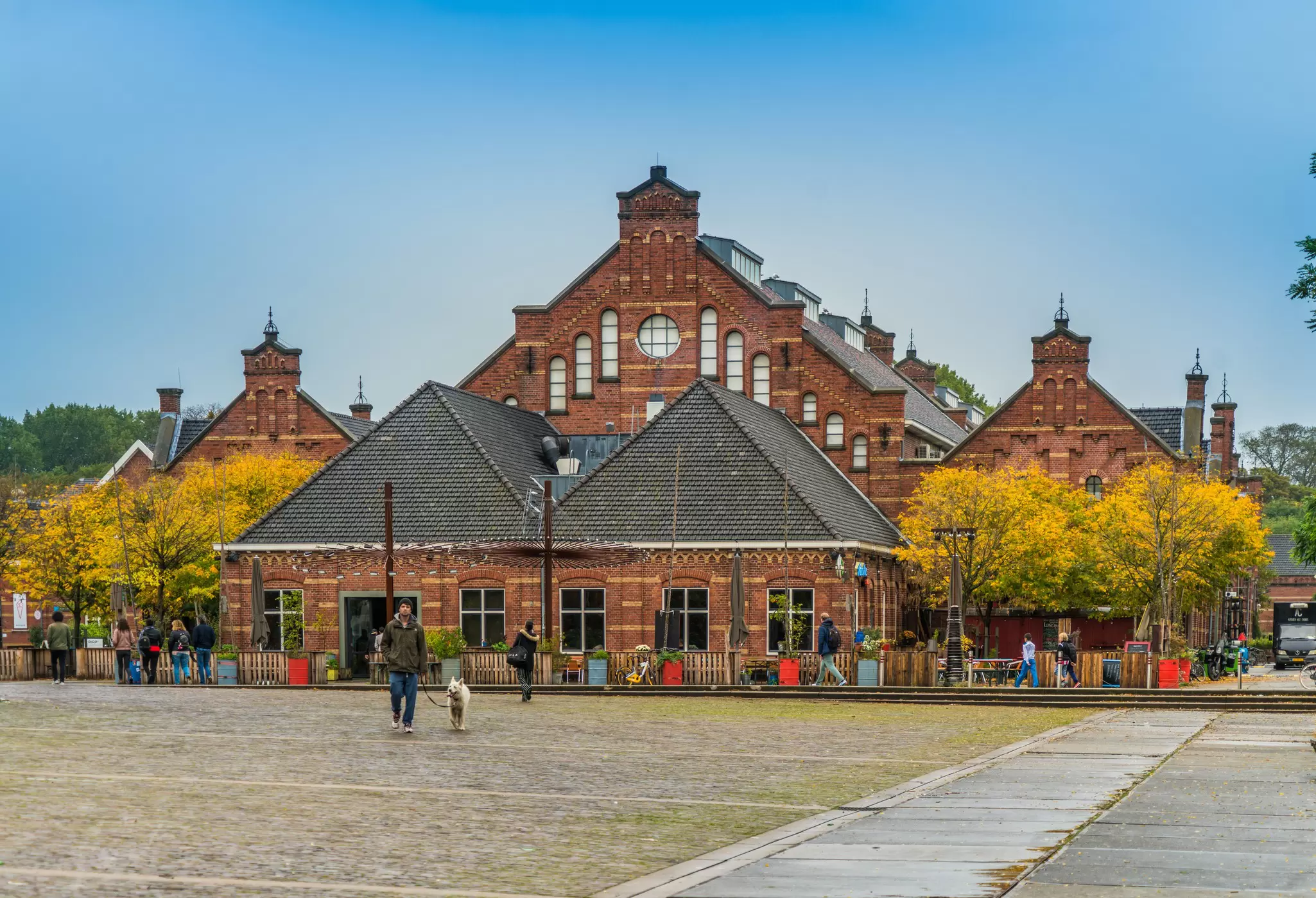 Restored red-brick warehouse buildings with stepped gables beside an autumn square, a man walking a white dog across the cobbles