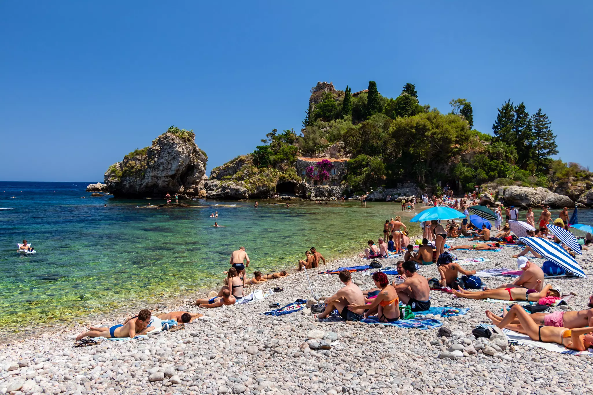 A crowd of people lie out on a rocky beach, with a small hill covered in trees in the background.