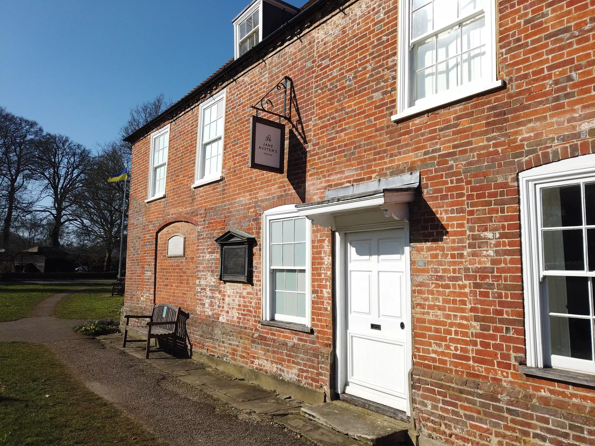 A side view of a two-story red brick house, with a white door in the center, white trim on the windows and a sign hanging from the second story.