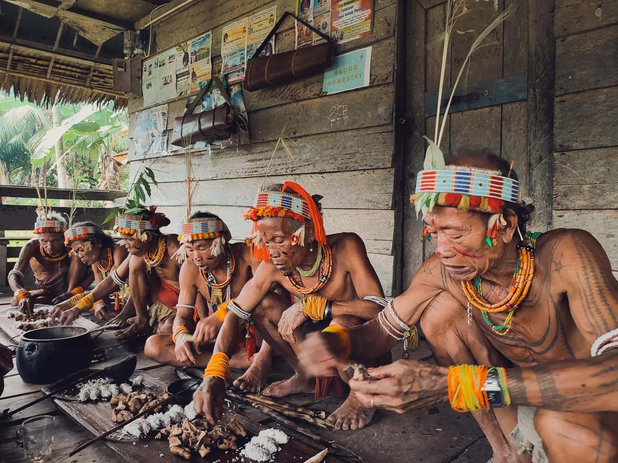 Six men wearing colorful headdresses, bracelets and necklaces squat low while eating a meal in a wooden shelter