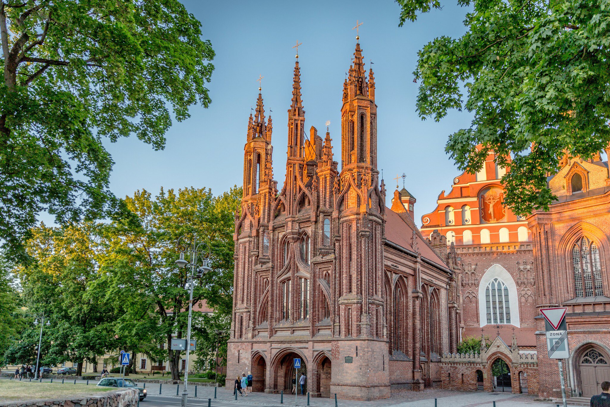 Three slender brick spires and pointed arches glow orange in evening light between leafy trees