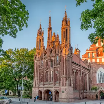 Three slender brick spires and pointed arches glow orange in evening light between leafy trees