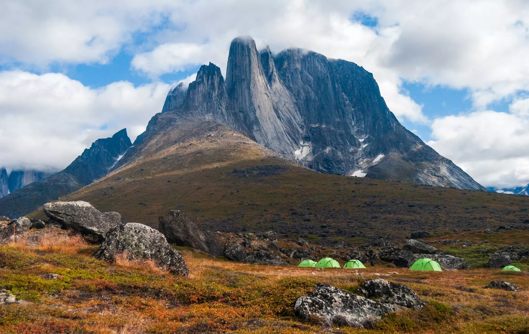 Five neon green tents are set up in scrubby vegetation at the base of a rocky mountain in Greenland.
