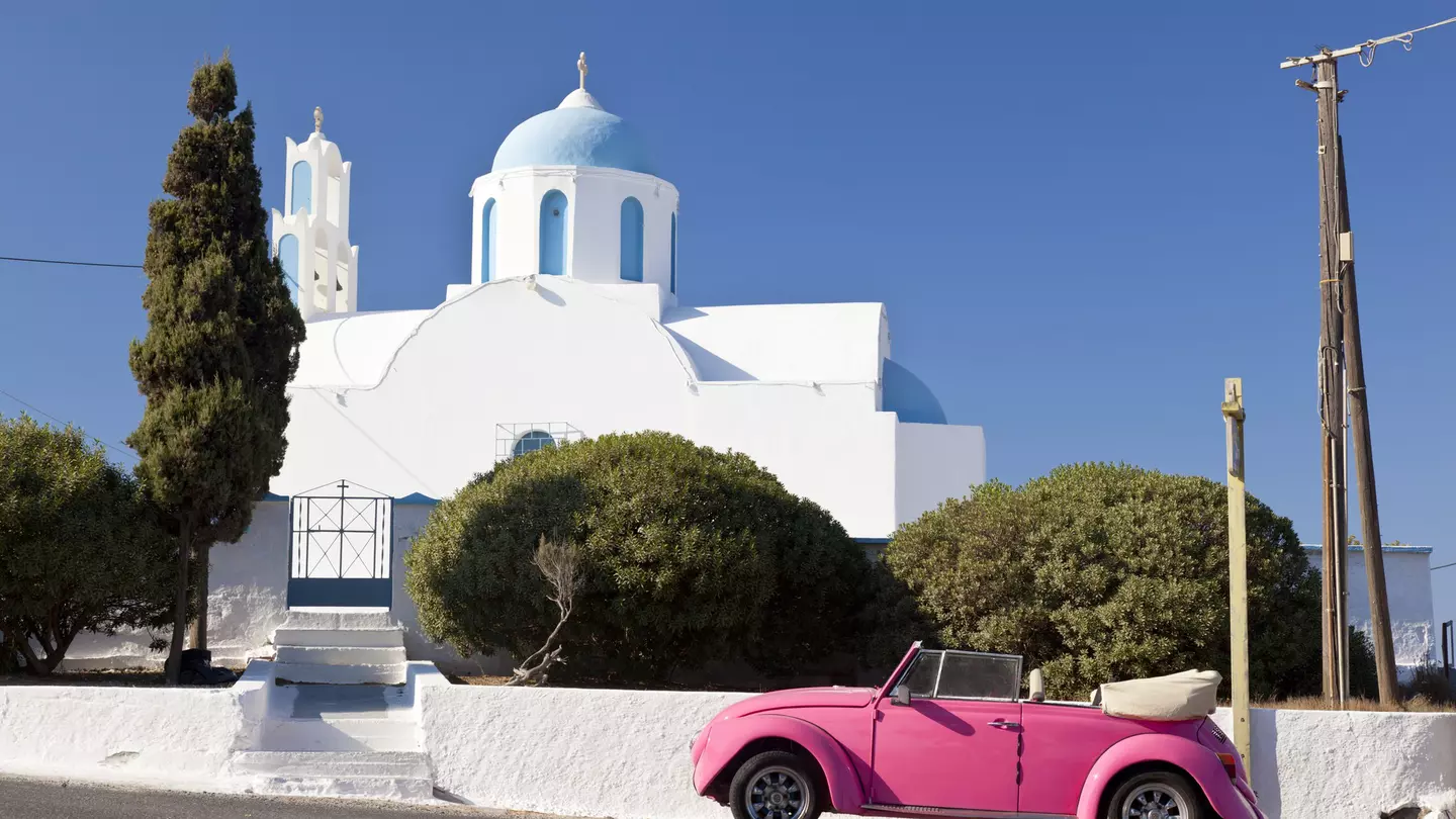 A pink Volkswagen Beetle in front of a chapel on Santorini, Greece