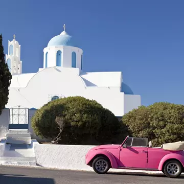 A pink Volkswagen Beetle in front of a chapel on Santorini, Greece