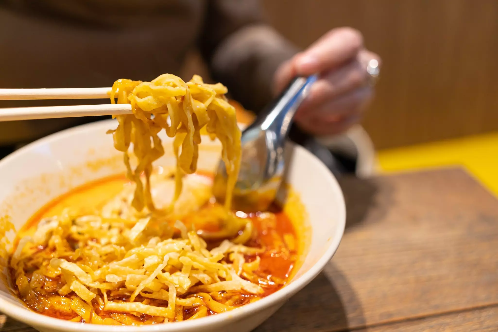 A close-up shot of a diner eating khao soi, fried noodles in creamy soup, with chopsticks and a metal spoon.