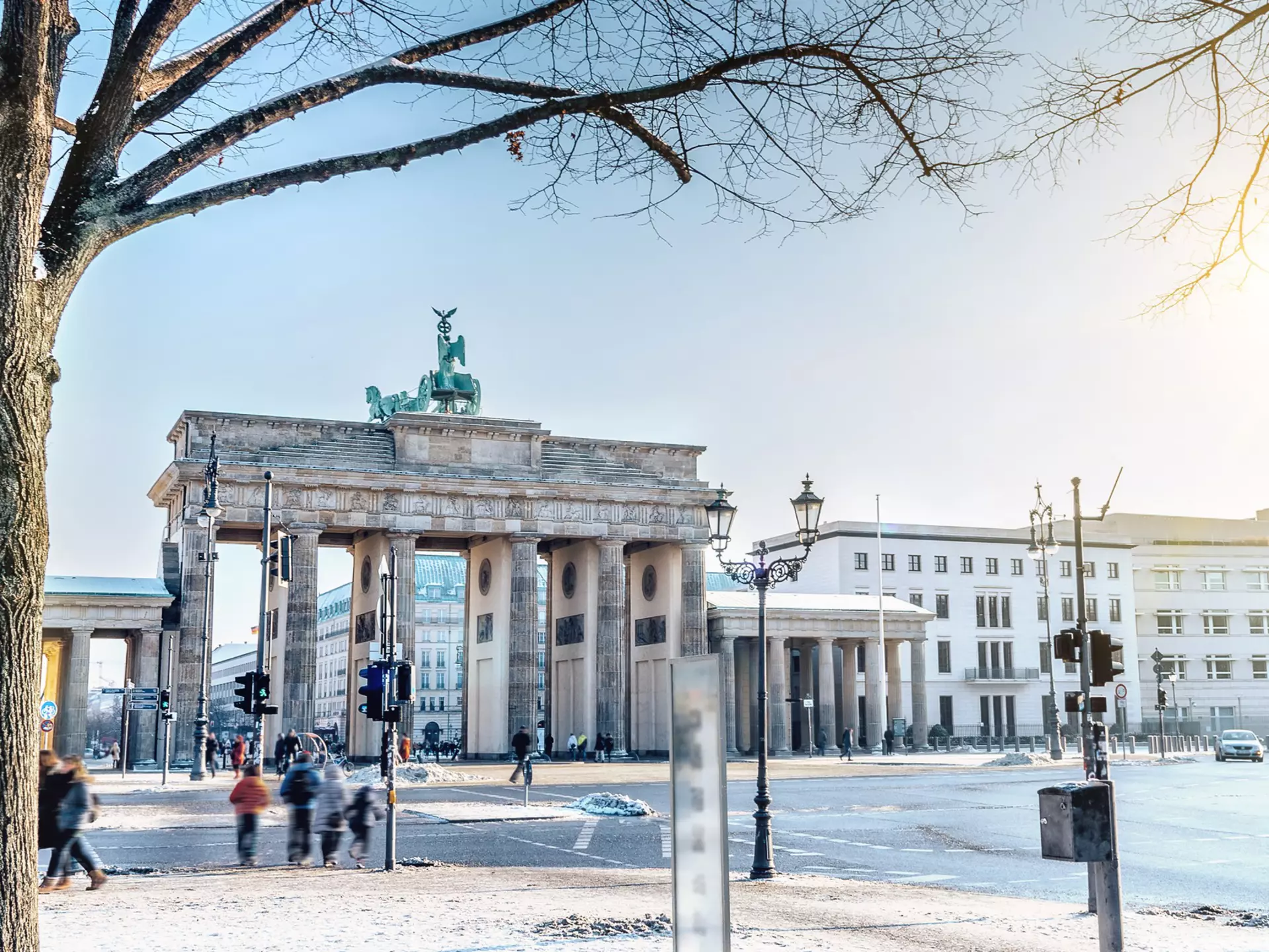 Out-of-focus people walk along a winter city street with a large stone gate along the roadway on a sunny day.