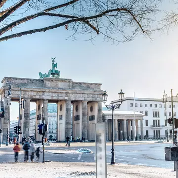 Out-of-focus people walk along a winter city street with a large stone gate along the roadway on a sunny day.