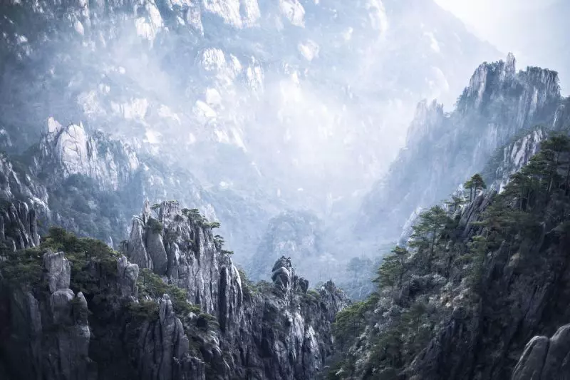 Rocky outcrops on a mountain are shrouded in mist, with the light creating a dramatic, painting-like effect.