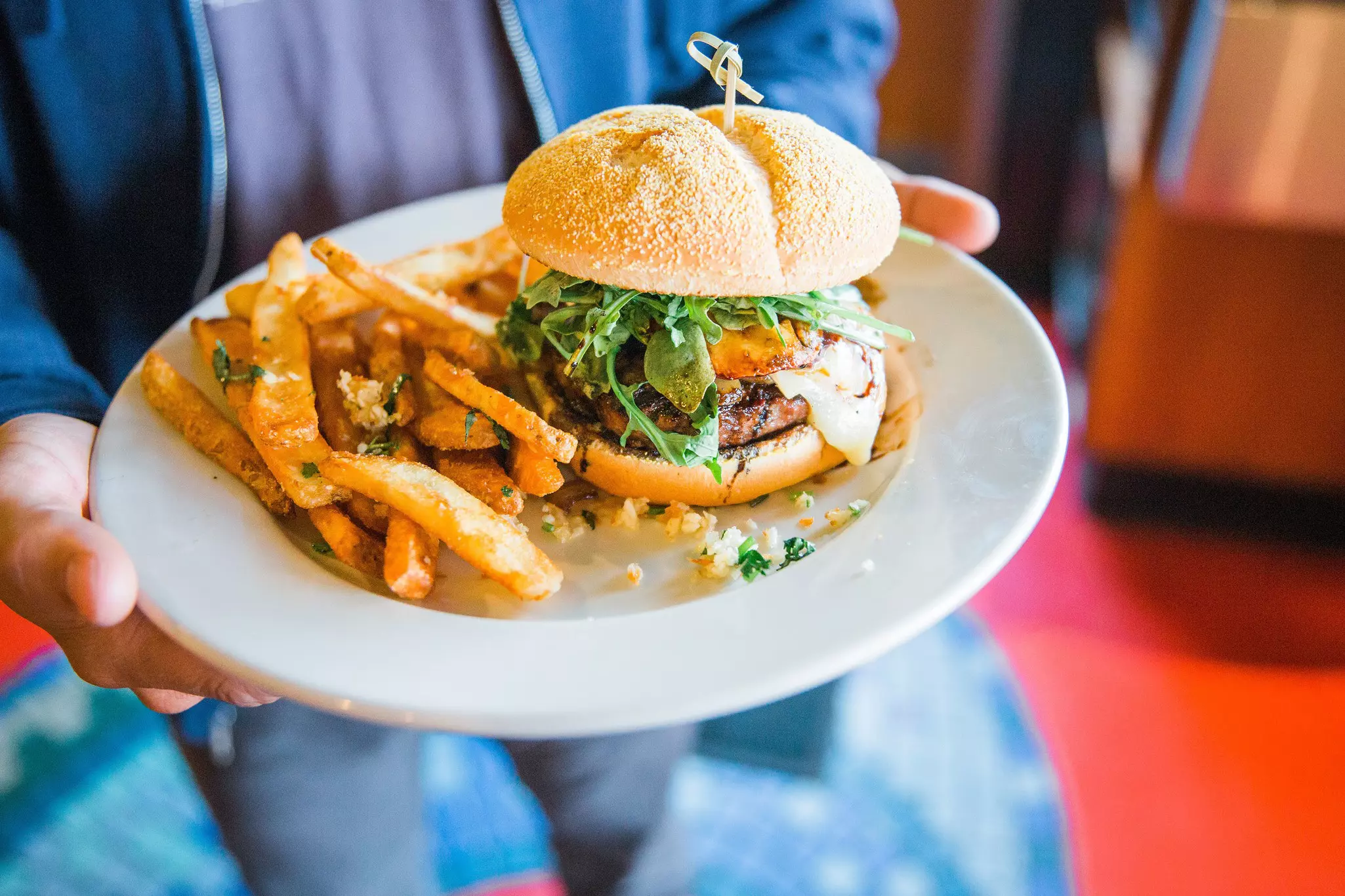Person in a blue sweatshirt holds a plate with a burger on a seseme seed bun topped with lettuce and accompanied by a side of fries