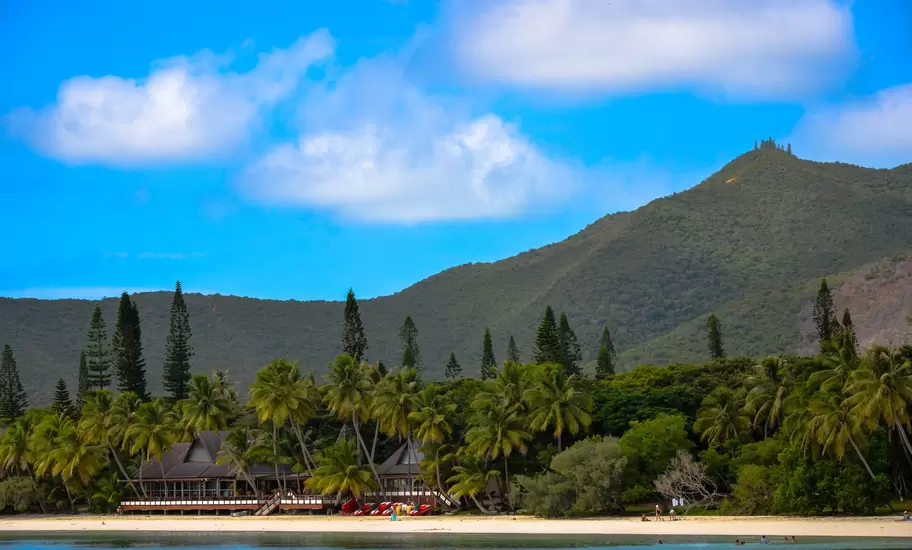 Isle of Pines beach in New Caledonia, with palms, tall columnar pines and a lodge beneath forested Pic N'ga