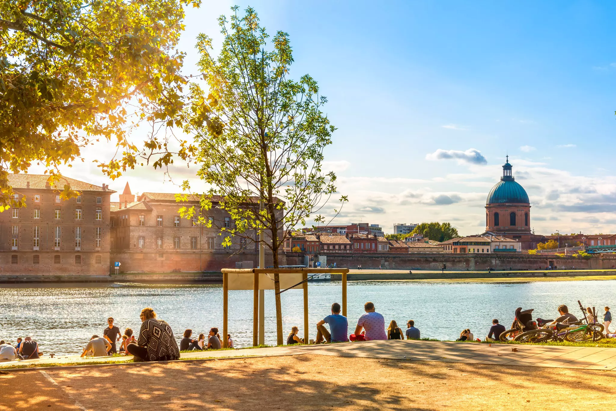 People sit on the edge of a riverbank watching the sun set. The light illuminates stone buildings and a church dome across the river.