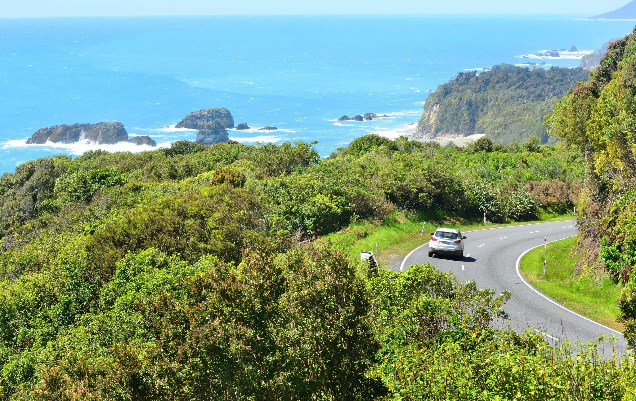 A single car drives on a two-lane road in New Zealand that winds along a bend with lush greenery on each side and the blue ocean to the left.
