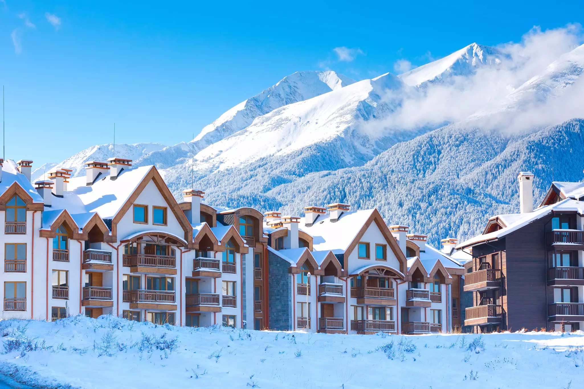 Townhomes with balconies and snow-covered peaked roofs are in front of snowcapped mountains in Bulgaria.