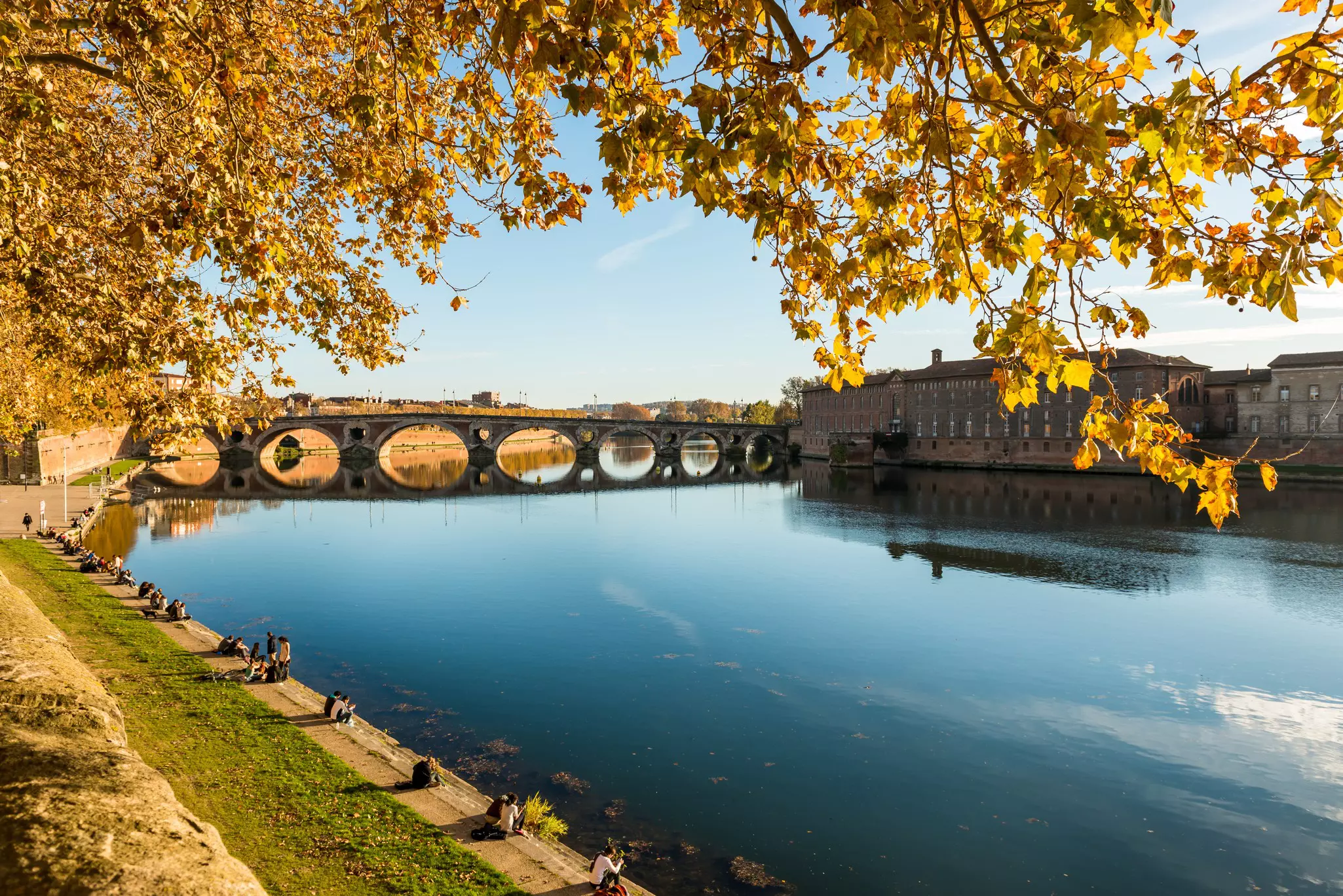 A riverside city with an arched bridge bathed in the golden light of autumn sunshine. People sit together in groups along the river bank.