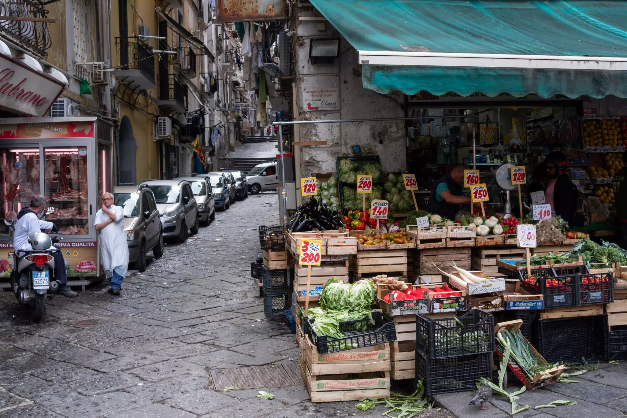 A stall in a city plaza sells fresh vegetables. Cars are parked along a narrow street intersecting with the plaza.