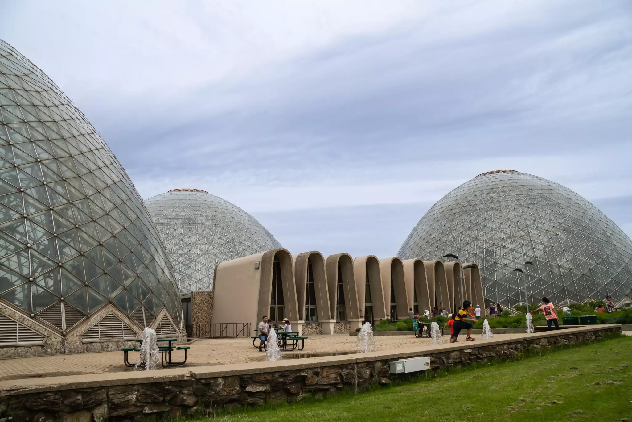 People relaxing at benches and along a stone walkway with large building-sized glass domes in the background on a cloudy day.