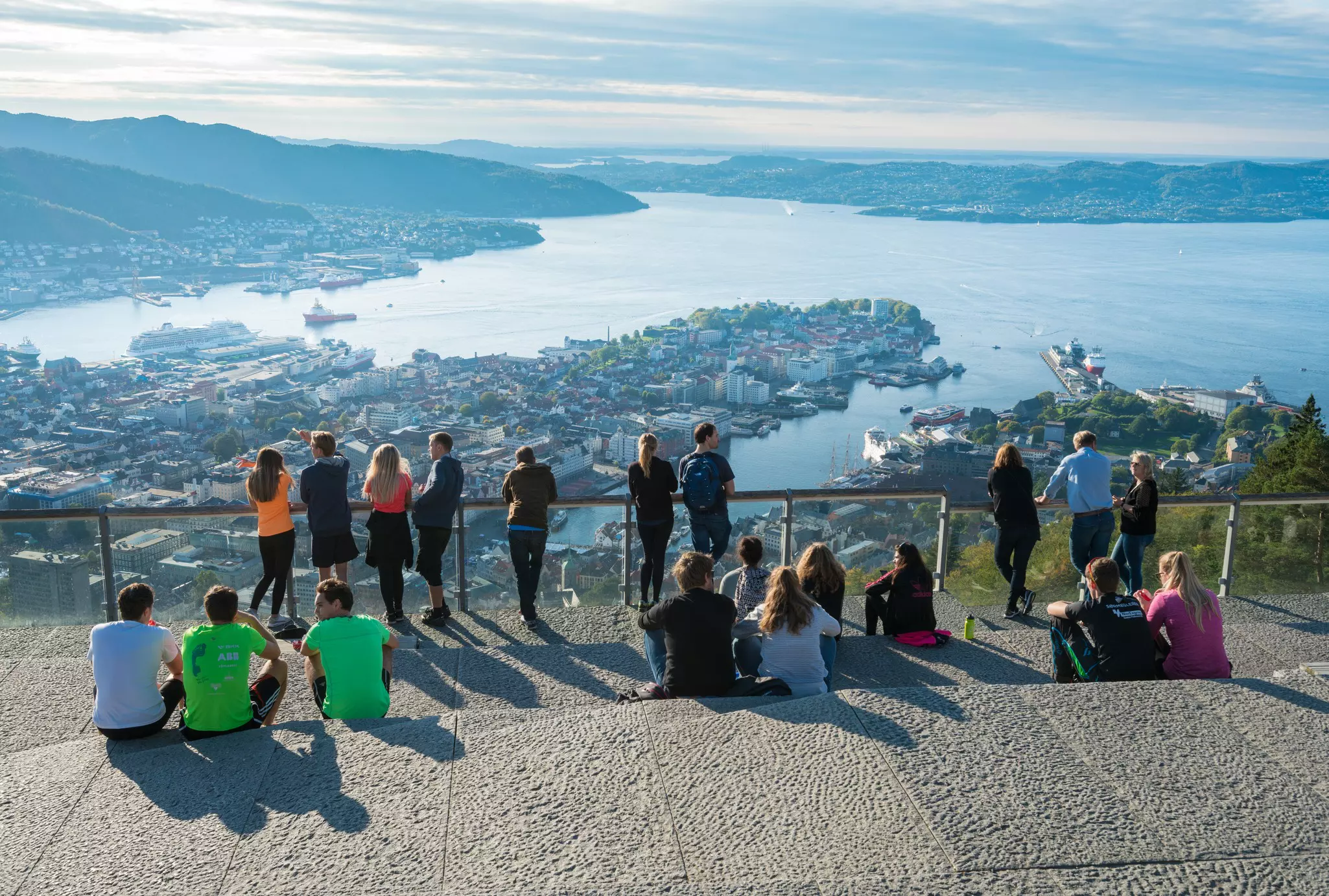 People stand at a viewpoint barrier looking down towards a waterside city