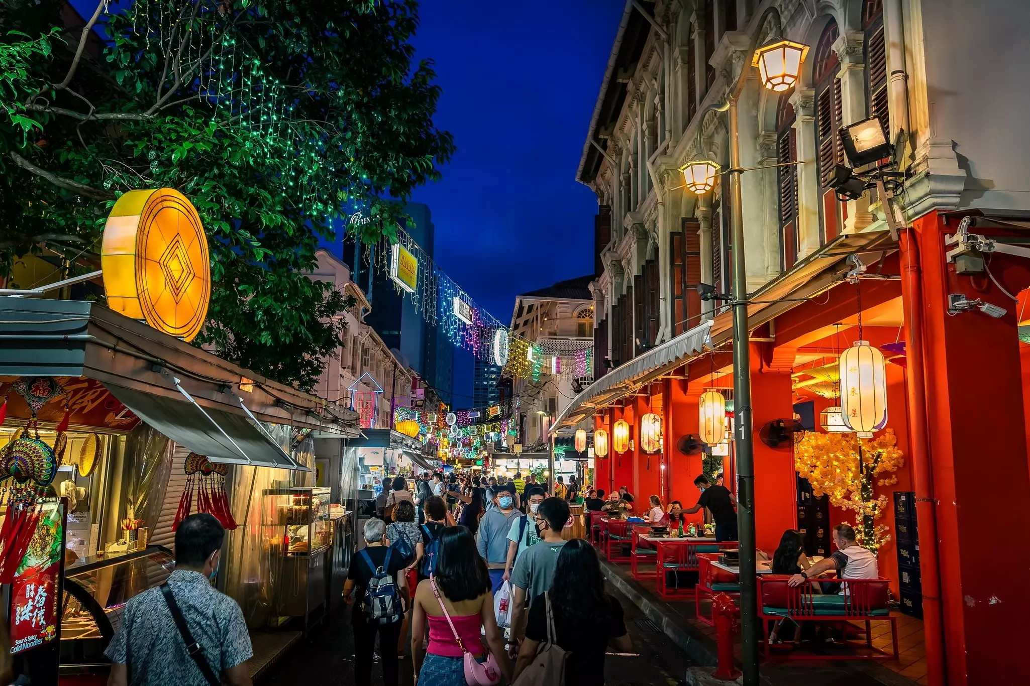 People walk down a city street at night, past lanterns and colorfully lit storefronts.