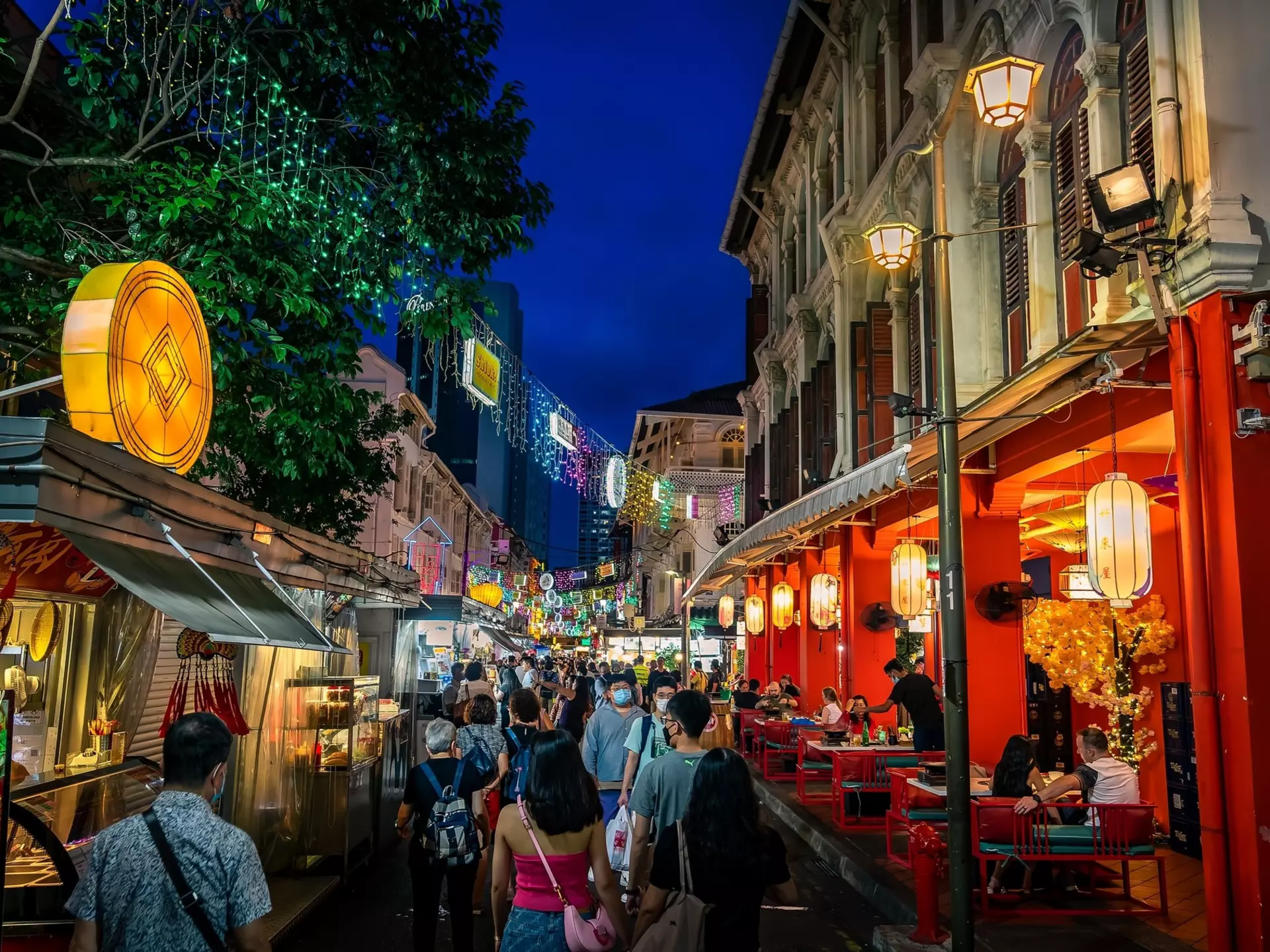 People walk down a city street at night, past lanterns and colorfully lit storefronts.