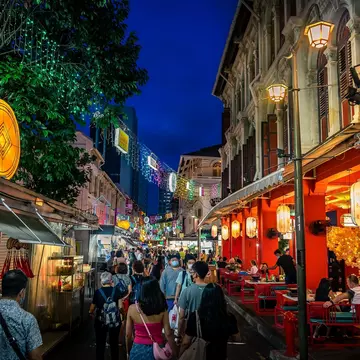 People walk down a city street at night, past lanterns and colorfully lit storefronts.