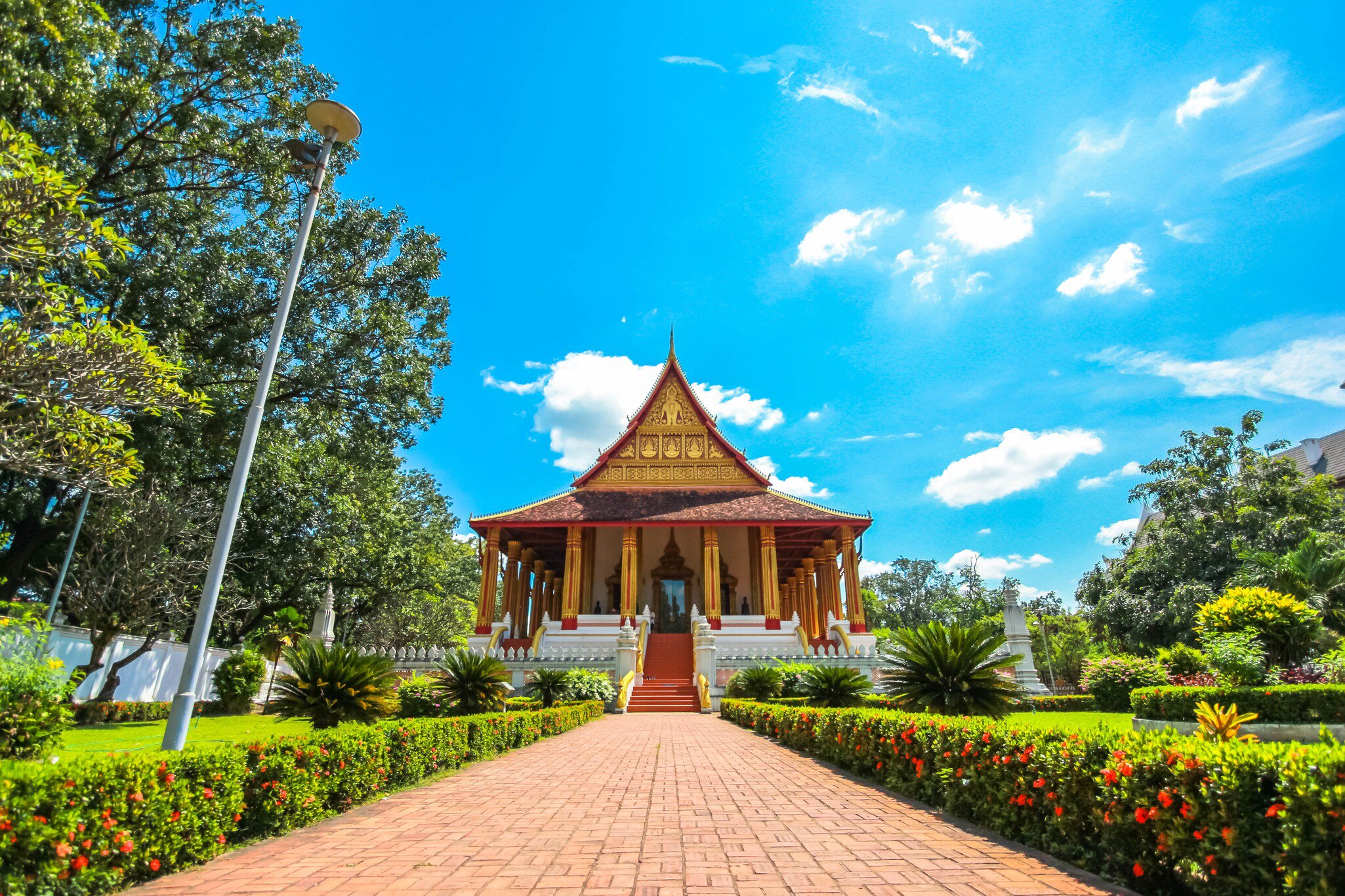 Red-roofed Lao temple hall with gilded columns and a red staircase at the end of a brick garden path.