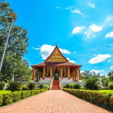 Red-roofed Lao temple hall with gilded columns and a red staircase at the end of a brick garden path.