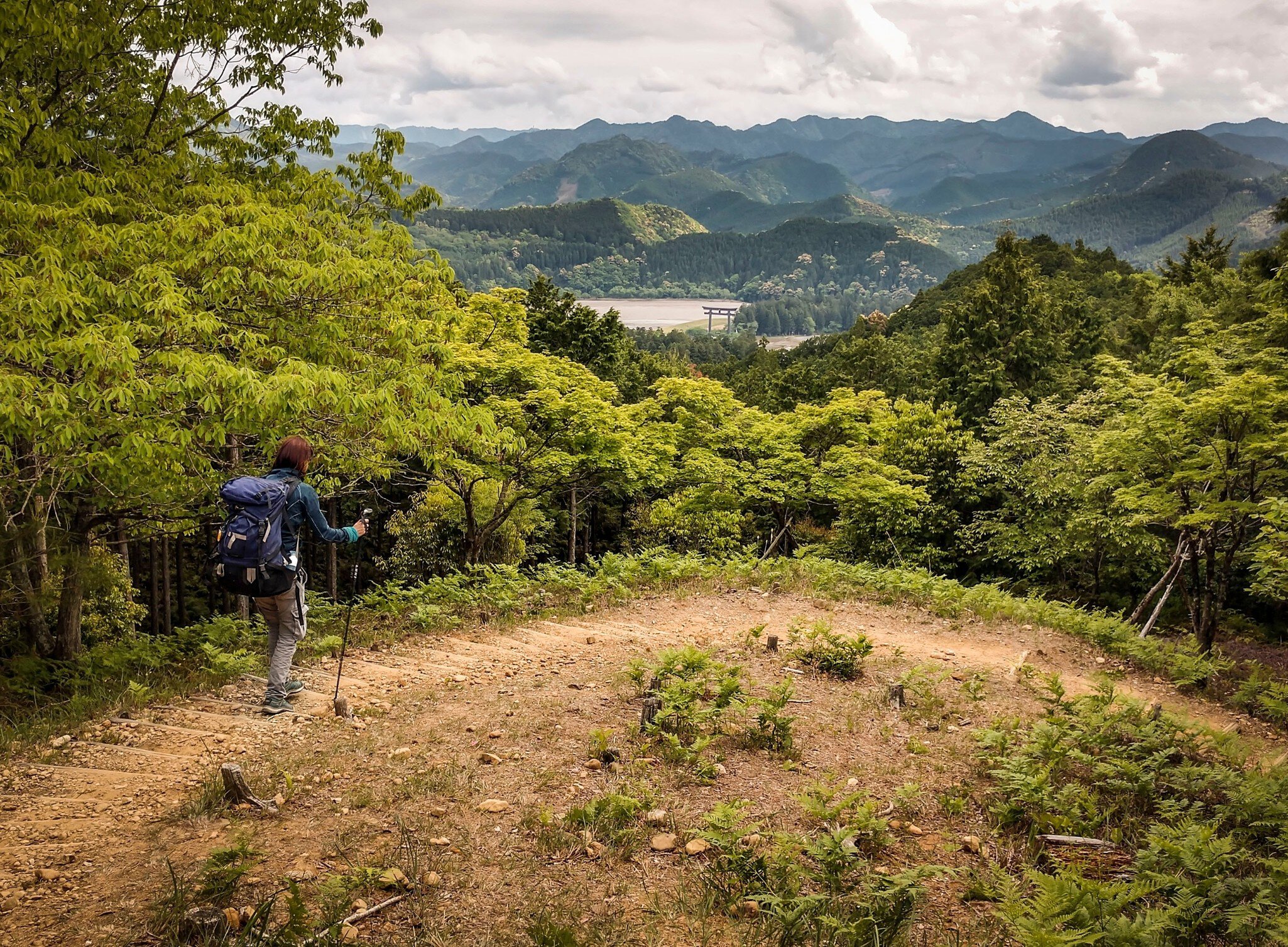A hiker on a dirt path in a leafy elevated location; a body of water is visible below, with a Japanese gate.