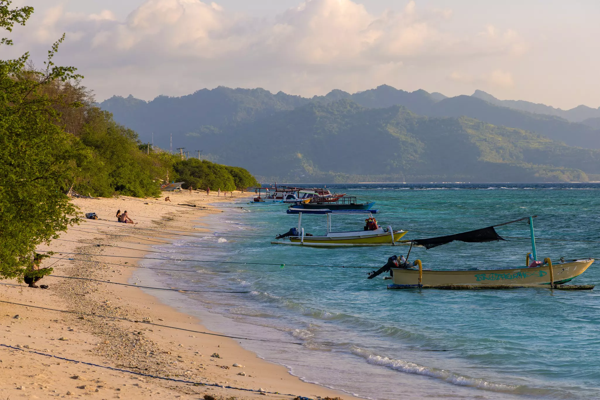 Fishing boats are moored off a narrow beach on a tropical island. Mountains are seen in the distance.
