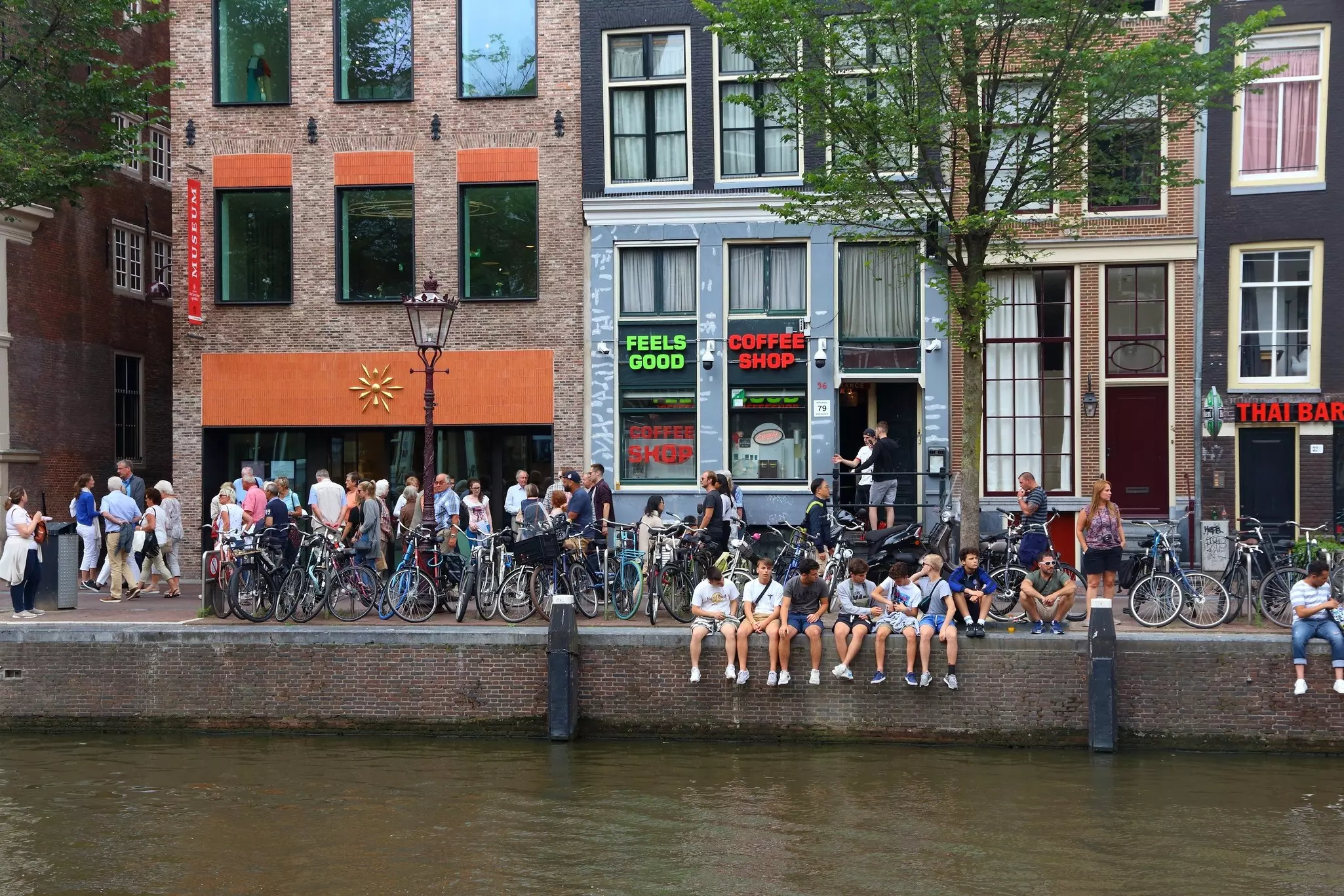 The exterior of Coffeeshop Amsterdam which is painted a dark khaki. The large windows feature the shops logos and are obscured by blinds. Two women are leaning against the window ledge outside the shop.