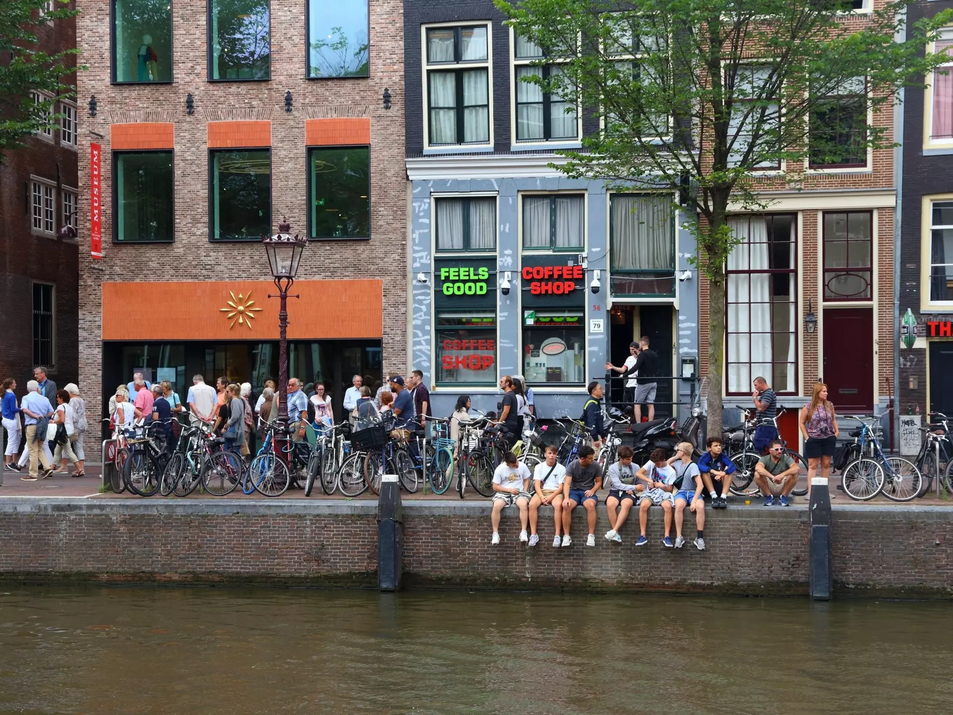 The exterior of Coffeeshop Amsterdam which is painted a dark khaki. The large windows feature the shops logos and are obscured by blinds. Two women are leaning against the window ledge outside the shop.
