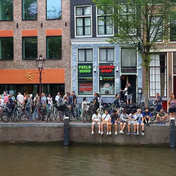 The exterior of Coffeeshop Amsterdam which is painted a dark khaki. The large windows feature the shops logos and are obscured by blinds. Two women are leaning against the window ledge outside the shop.