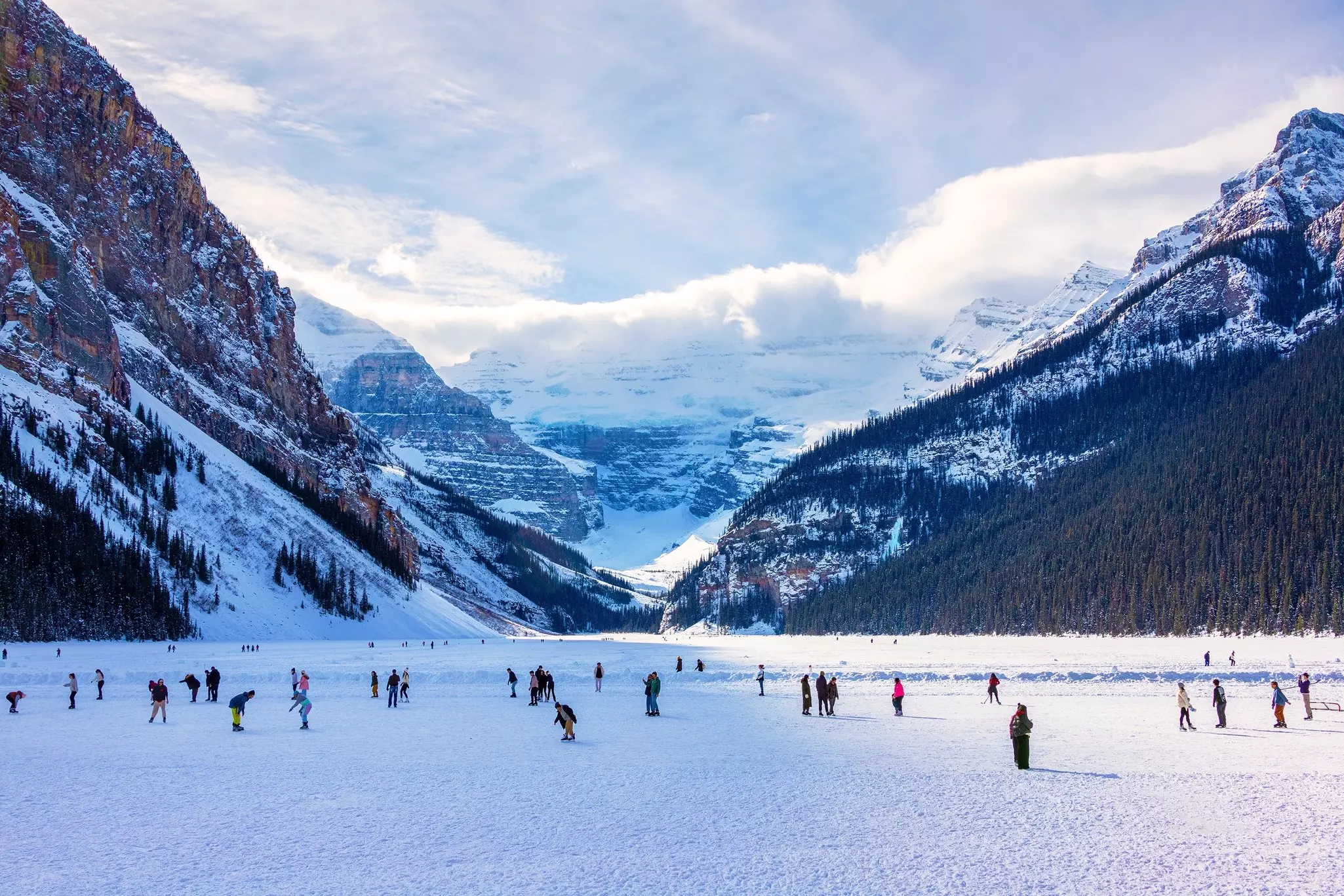 People ice skate on a frozen lake surrounded by snowcapped mountains.