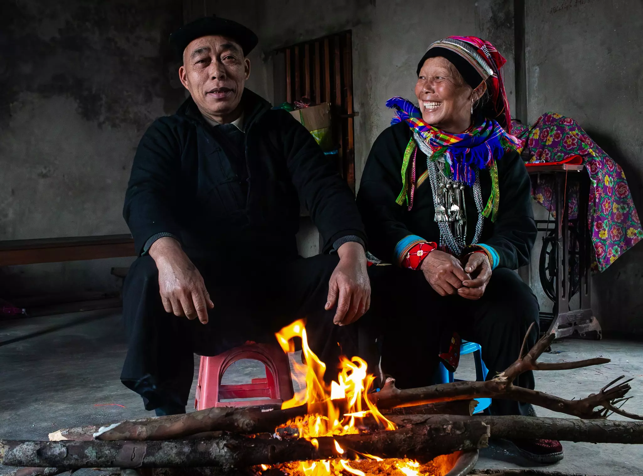 A man and woman sit on stools in front of a small fire. She is wearing colorful woven accessories.