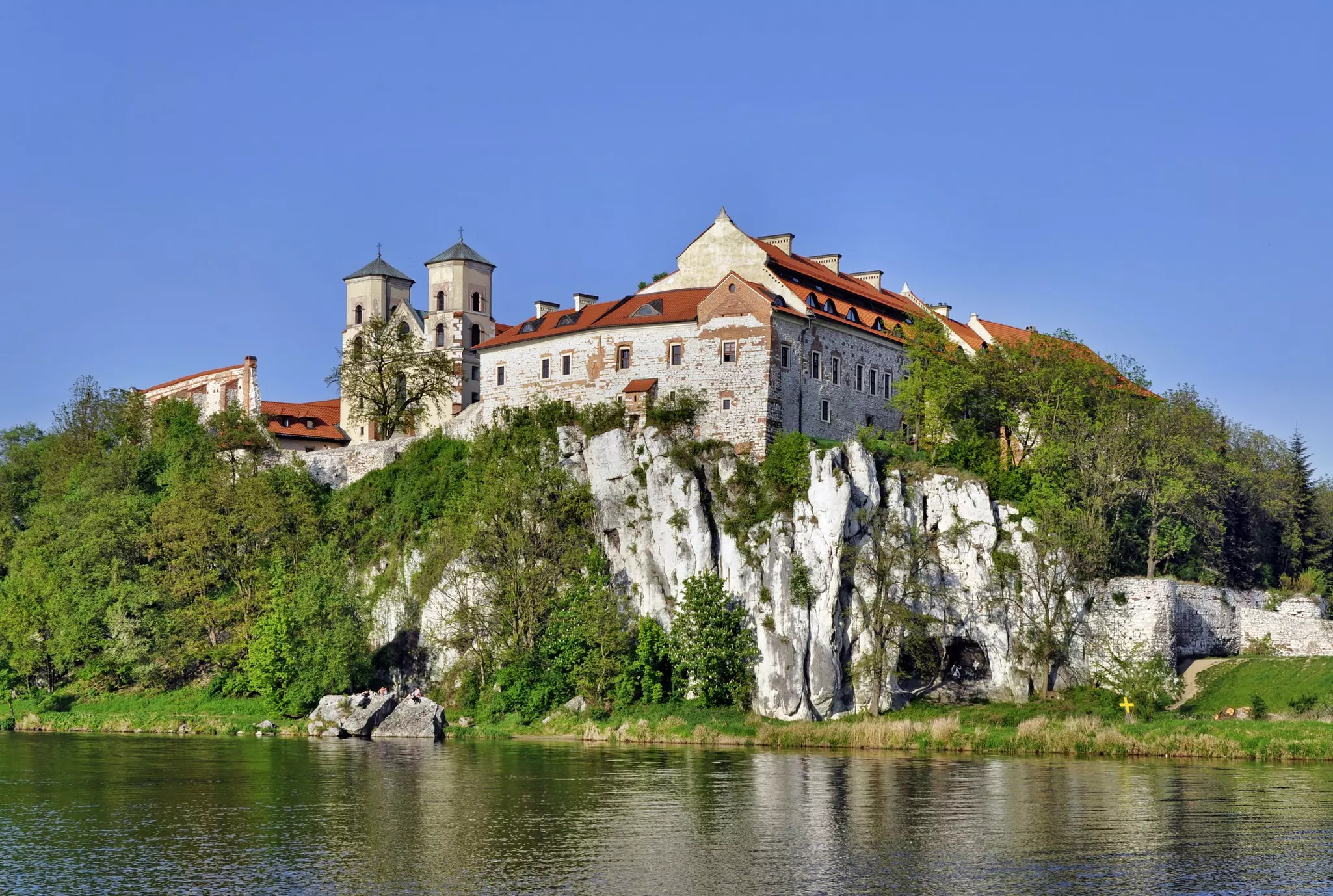 A stone building with a red roof on a rocky hill above a river with grass and trees on the shore.