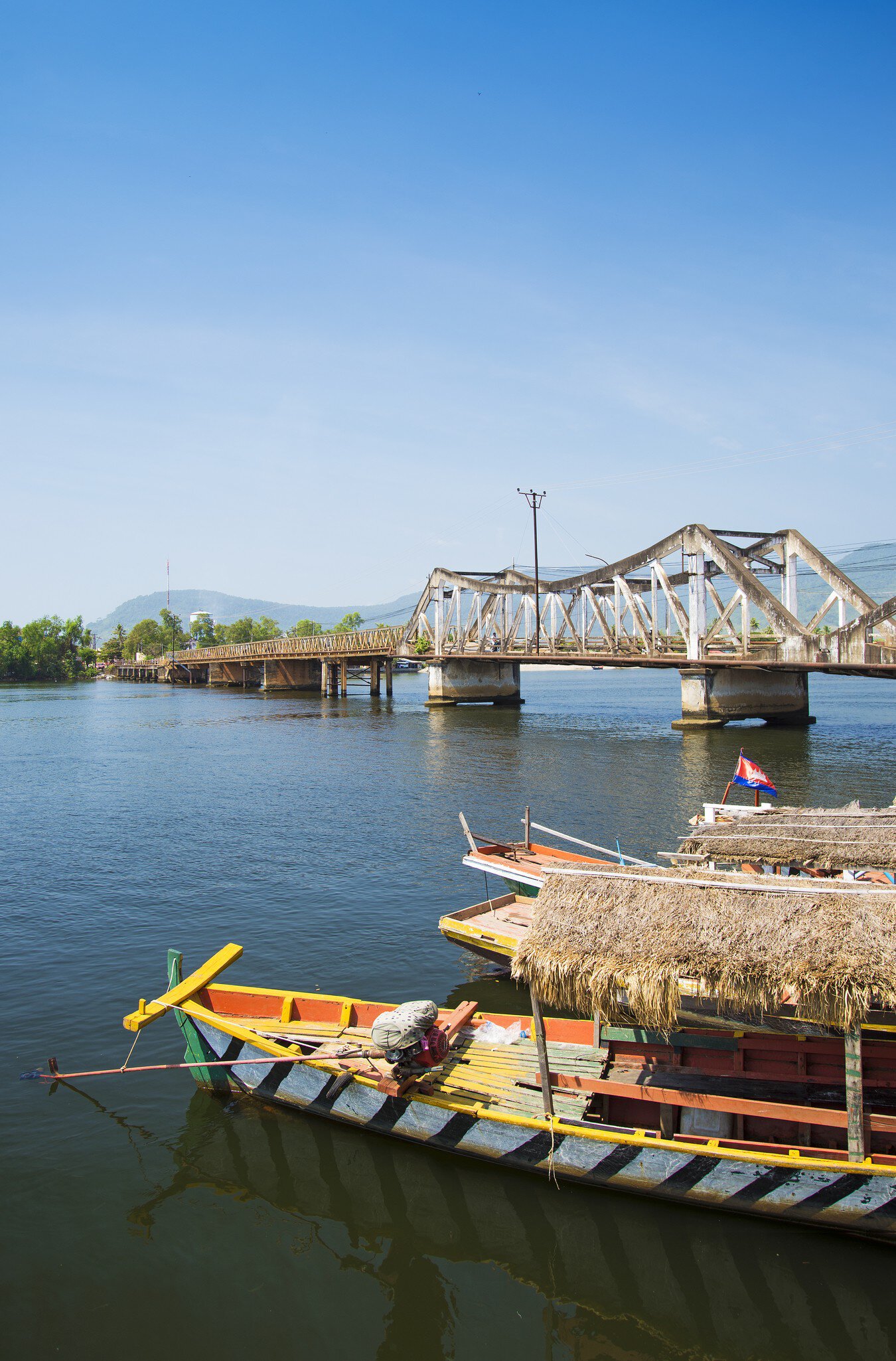 Painted wooden boats with thatched roofs moored on a river in front of a steel truss bridge, Kampot, Cambodia.