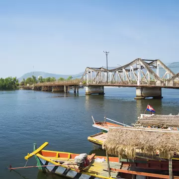 Painted wooden boats with thatched roofs moored on a river in front of a steel truss bridge, Kampot, Cambodia.