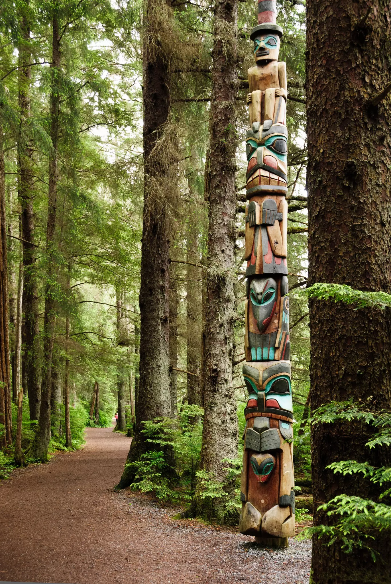 A large wooden carved totem pole at the edge of a gravel path through dense woodland.