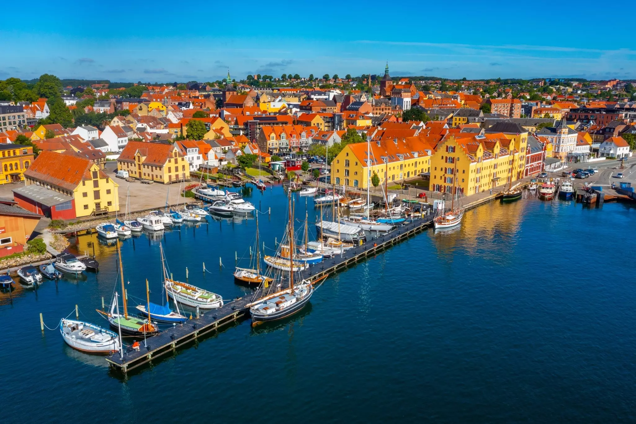 An aerial view of a port town. Sailboats are moored at a long dock, while yellow-painted buildings lie just offshore.