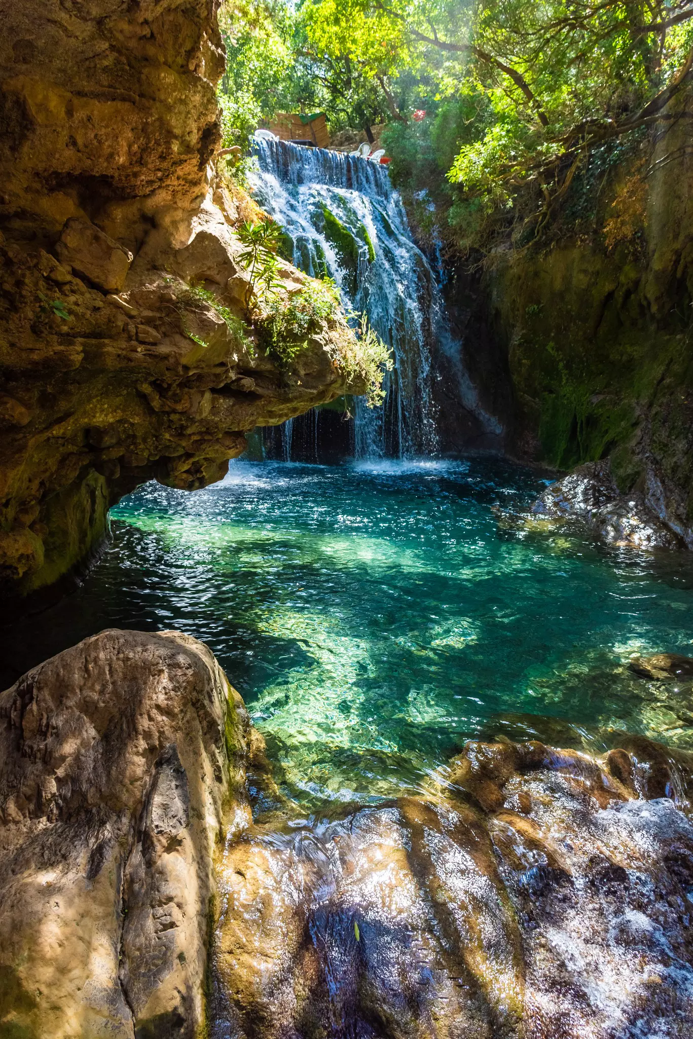 A waterfall pours into a deep turquoise pool, with rocks in the foreground. 